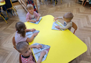 Zdjęcie przedstawia dzieci siedzące przy stole, składające gazetę.