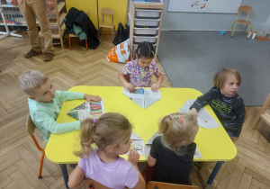 Na zdjęciu widać dzieci siedzące przy stoliku, które słuchają jak mają wykonać czapkę z papieru