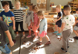 Na zdjęciu widzimy grupkę dzieci, które maszerują pomiędzy swoimi pracami stworzonymi z jesiennych materiałów przyrodniczych.