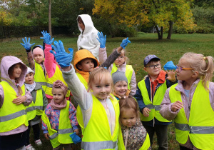 Na zdjęciu widzimy grupkę dzieci, które uczestniczą w akcji "Sprżatamy dla Polski". Każde dziecko ma niebieską rękawiczkę na jednej z dłoni.