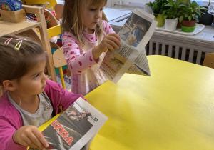 Dzieci siedzą przy stoliku i składają czapeczki z gazety.