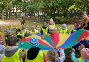 Zdjęcie przedstawia dzieci i nauczycielkę bawiących się z wykorzystaniem chusty animacyjnej. Grupa znajduje się w parku.