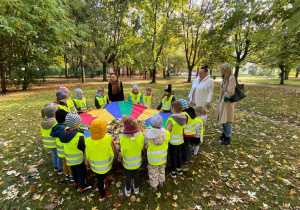 Zdjęcie przedstawia grupę przedszkolną w parku. Dzieci zgromadzone są wokół chusty animacyjnej.