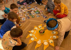 Zdjęcie przedstawia dzieci siedzące w kole. Na środku na podłodzie leży urodzinowe słoneczko Montessori oraz fotografie. Chłopiec w niebieskiej koronie wkłada koraliki do miseczek.