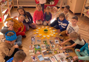 Zdjęcie przedstawia dzieci siedzące w kole. W środku na podłodze leżą zdjęcia oraz urodzinowe słoneczko Montessori.