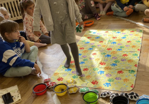 Dzieci siedzą w kole, a dziewczynka idzie po wybranym kolorze psich łapek na dywaniku.