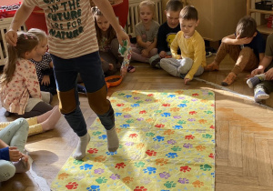 Dzieci siedzą w kole, a chłopiec idzie po wybranym kolorze psich łapek na dywaniku.