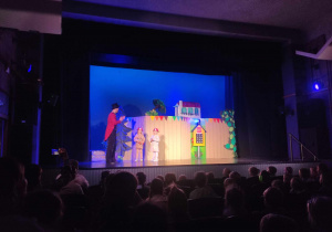 Zdjęcie przedstawia scenę teatralną, na której stoi aktor i dwie dziewczynki. W tle jest dekoracja.