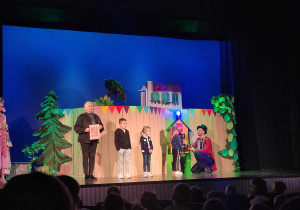 Zdjęcie przedstawia scenę teatralną na której stoi dwoje aktorów i trójka dzieci.