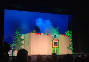 Zdjęcie przedstawia scenę teatralną. Na scenie jest dekoracja- płot, za nim domek z którego wydobywa się dym. Przed płotem jest budka z telefonem.