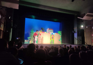 Zdjęcie przedstawia scenę teatralną, na której stoi aktor i dwie dziewczynki. W tle jest dekoracja.
