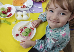 Na zdjęciu widzimy uśmiechniętą dziewczynkę, która trzyma przed sobą na talerzyku kolorową kanapkę.
