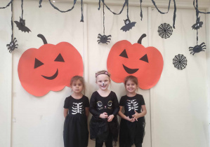Zdjęcie przedstawia trzy dziewczynki w strojach na tle halloweenowej dekoracji.