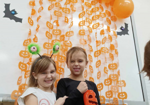 Na zdjęciu widać dwie dziewczynki stojące przy tablicy ozdobionej dyniami i pomarańczowymi balonami.