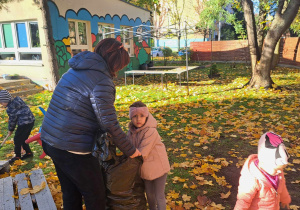 Zdjęcie przedstawia nauczyciela z dziećmi, który pomaga zbierać liście w ogrodzie