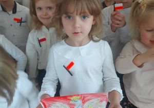 Na zdjęciu widać grupę dzieci, przed którymi soi dziewczynka i trzyma pokolorowany obrazek godła polskiego
