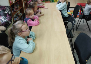 Na zdjęciu widzimy grupkę dzieci, które czekają na zajęcia czytelnicze w Bibliotece. Dzieci siedzą przy ławkach.