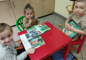 Na zdjęciu widzimy trójkę dzieci, które siedzą przy czerwonym stoliku i oglądają książki.