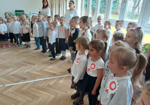 Na zdjęciu widzimy grupkę dzieci i nauczycielki, które śpiewają Hymn Polski. Wszystkie dzieci i nauczycielki ubrani są na galowo.