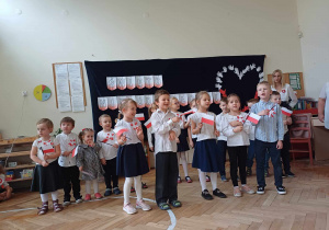Na zdjęciu widzimy dzieci z grupy I, które śpiewają piosenkę patriotyczną "Polska - moją ojczyzną kochaną". Wszystkie dzieci ubrane są na galowo i trzymają w ręku flagę Polski.