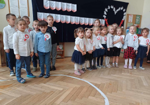Zdjęcie przedstawia dzieci ustawione tak, że z lewej strony stoją chłopcy a z prawej dziewczynki. Dzieci śpiewają piosenkę. W tle znajduje się dekoracja z okazji Święta Niepodległości.