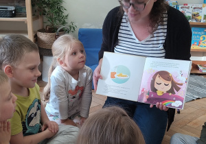 Mama czyta dzieciom książkę, a dzieci siedzą na podłodze i słuchają.