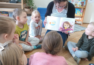 Mama czyta dzieciom książkę, a dzieci siedzą na podłodze i słuchają.