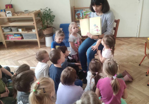 Zdjęcie przedstawia kobietę siedzącą na krześle, która czyta dzieciom książkę. Dzieci siedzą na podłodze i słuchają czytanego utworu.