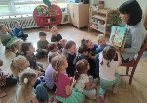 Zdjęcie przedstawia kobietę siedzącą na krześle, która czyta dzieciom książkę. Dzieci siedzą na podłodze i słuchają czytanego utworu.