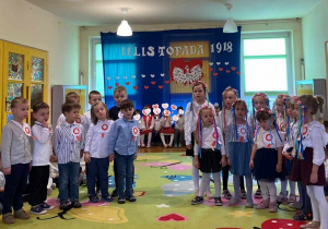 Zdjęcie przedstawia dzieci ustawione tak, że z lewej strony stoją chłopcy a z prawej dziewczynki. Dzieci śpiewają piosenkę. W tle znajduje się dekoracja z okazji 11 listopada.