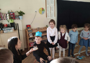 Zdjęcie przedstawia chłopca w niebieskiej koronie, który zdmuchuje świeczki z tortu trzymanego przez nauczycielkę. Czynności przyglądają się dzieci zgromadzone wokół.