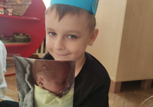 Zdjęcie przedstawia chłopca w niebieskiej koronie, który pokazuje fotografię przedstawiającego go jako noworodka.