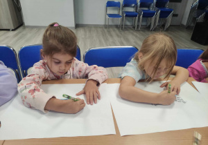 Zdjęcie przedstawia dzieci siedzące przy stole. Dzieci wykonują prace plastyczne na dużych arkuszach z wykorzystaniem kredek.