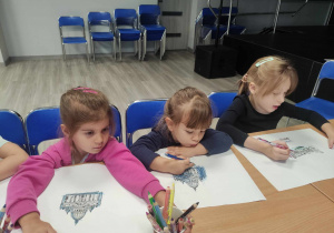 Zdjęcie przedstawia dzieci siedzące przy stole. Dzieci wykonują prace plastyczne na dużych arkuszach z wykorzystaniem kredek.