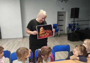 Zdjęcie przedstawia dzieci siedzące przy stole. Dzieci słuchają kobiety, która pokazuje im dużą fotografię przedstawiającą koloseum.