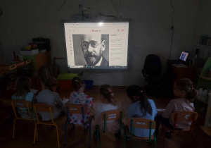 Zdjecie przedstawia dzieci siedzące przed tablicą na której jest postać Janusza Korczaka