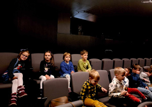 Zdjęcie przedstawia dzieci siedzące na fotelach w sali teatralnej.