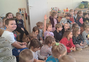 Zdjęcie przedstawia dziewczynkę siedzącą wśród innych dzieci, podnoszącą rękę do odpowiedzi.