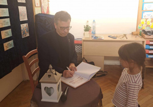 Zdjęcie przedstawia mężczyznę podpisującego książkę, dla dziewczynki stojącej obok.
