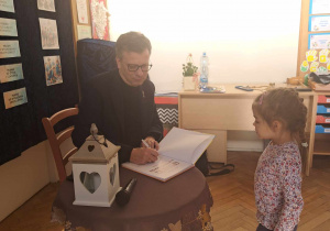 Zdjęcie przedstawia mężczyznę podpisującego książkę, dla dziewczynki stojącej obok.