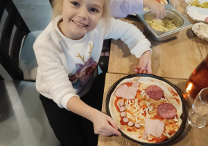 Dziewczynka pokazuje talerz na którym ma ułożoną buźkę z różnych składników pizzy.
