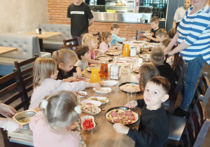Dzieci siedzą przy wspólnym stole i jedzą pizzę.