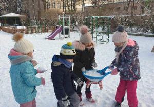 Grupka dzieci niesie taczkę ze śniegiem.