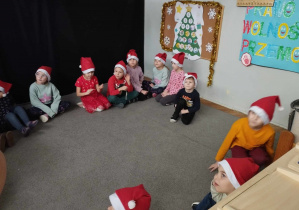 Na zdjęciu widzimy grupkę dzieci, które siedzą i słuchają pomocników - Pani Dyrektor i Pani Ewy. Wszystkie dzieci na głowach mają czapki mikołajowe.