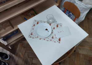 Na zdjęciu widzimy biały stolik, na którym położony jest talerzyk z okryszkami. Obok talerzyka stoi szklanka, która jest pusta. Obok szklanki znajduje się koperta z napisem "Grupa I".