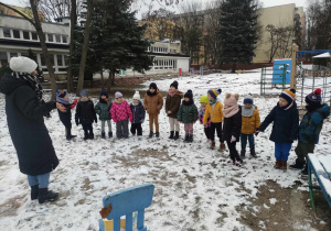 Zdjęcie przedstawia grupę dzieci stojących w rzędzie w ogrodzie przedszkolnym oraz nauczycielkę. W ogrodzie jest śnieg.