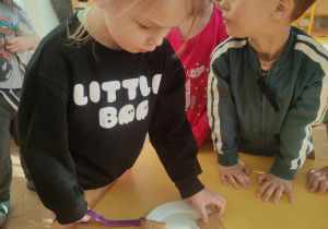 Zdjęcie przedstawia dziewczynkę kładącą na talerzyku ciasteczka. Czynności przyglądają się inne dzieci.