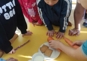 Zdjęcie przedstawia chłopca kładącego marchewki na talerzu. Czynności przyglądają się inne dzieci.