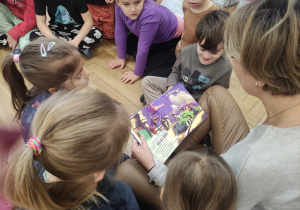 Zdjęcie przedstawia dzieci zgromadzone wokół kobiety, która czyta im książkę.