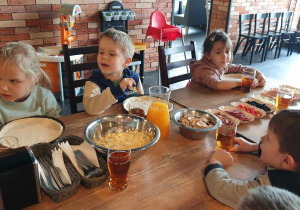 Zdjęcie przedstawia dzieci siedzących przy stole, słuchających wskazówek osoby dorosłej jak przygotować pizze.